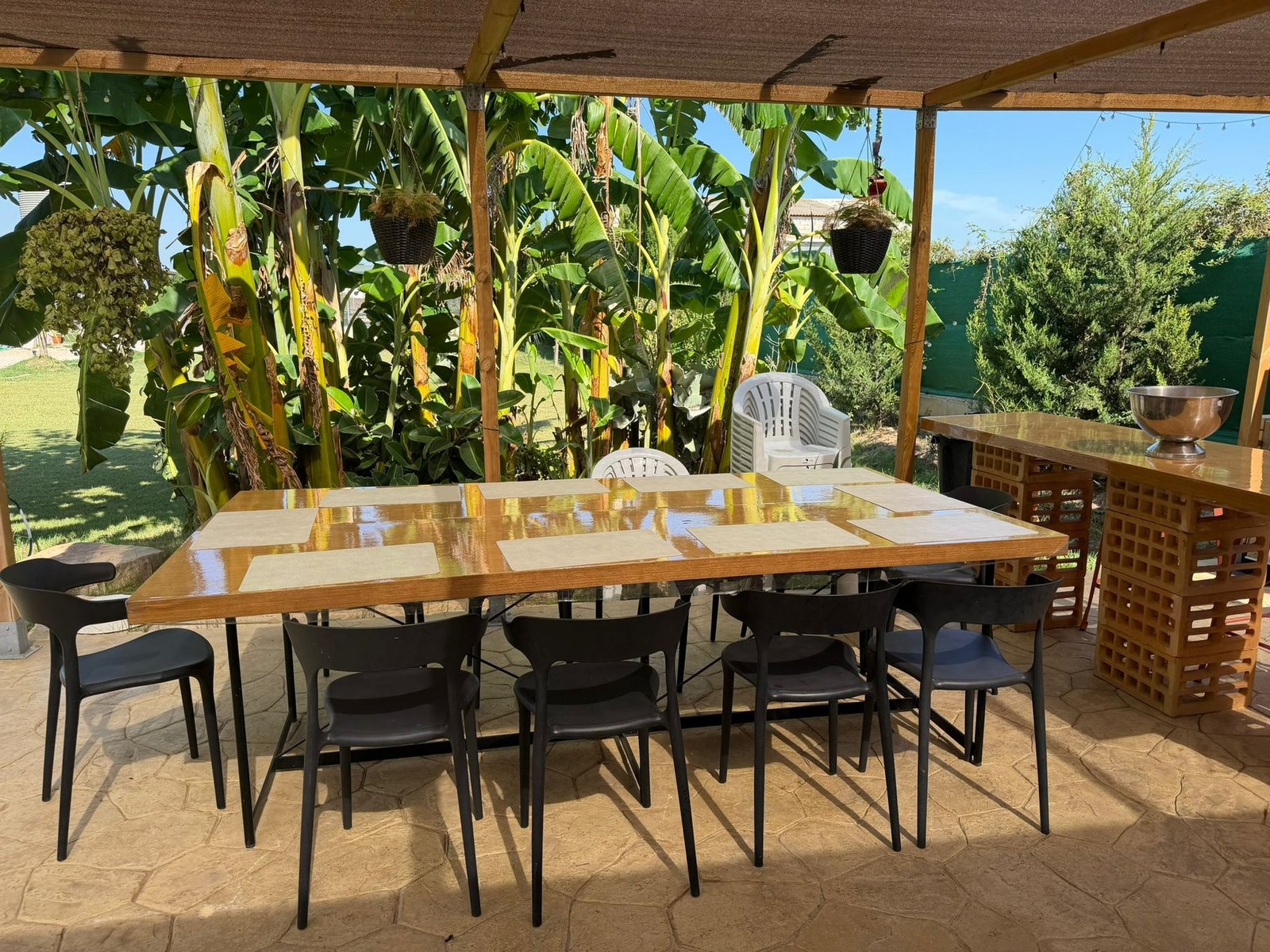 Mesa larga de comedor bajo la pérgola, entre plataneras, con barra de servicio