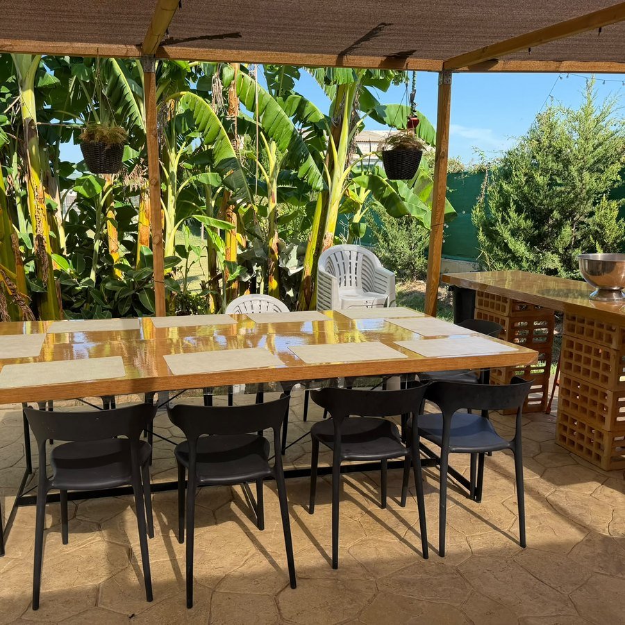 Mesa larga de comedor bajo la pérgola de madera, entre plataneras, en Knattur